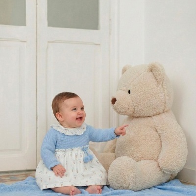 Bebé com vestido azul e branco sentada num tapete azul a apontar para um grande urso de peluche creme