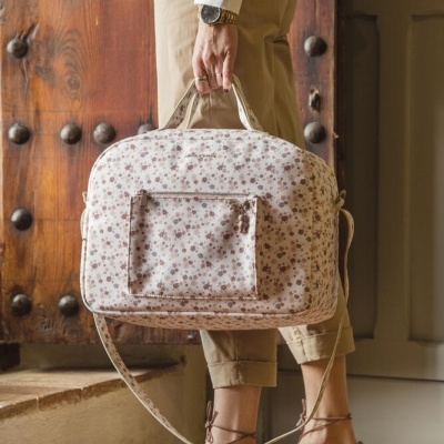 Mala floral de tecido com bolso externo e alças, segurada por pessoa junto a porta de madeira