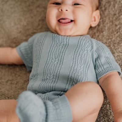 Bebé deitado, roupa de malha azul clara com padrão trançado.