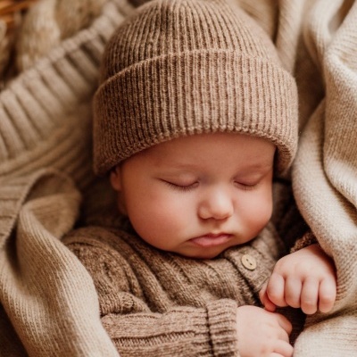 Bebé a dormir com gorro e camisola castanha e manta bege numa cesta.