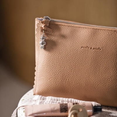 Carteira bege em pele sintética com fecho e pendente metálico, tubo de creme e frasco cosmético sobre superfície clara