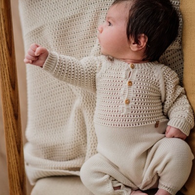 Bebé com fato de malha bege sentado numa poltrona com almofada e manta bege
