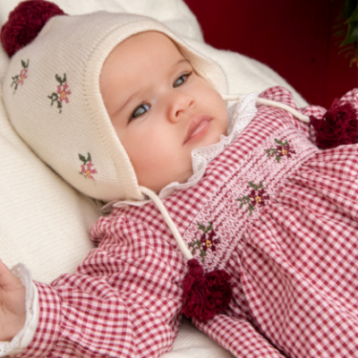 Bebé com vestido xadrez vermelho e branco e gorro creme com flores e pompons vermelhos