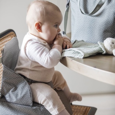 Bebé sentado em cadeira com almofada cinzenta de pontos brancos ao lado de mesa com saco e manta
