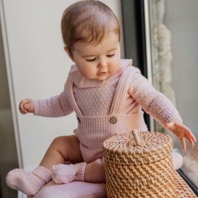 Bebé com roupa cor de rosa sentado junto a cesta de vime à beira da janela