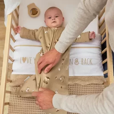 Bebé deitado em berço com pijama bege estampado e almofadas brancas com texto dourado