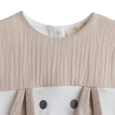 Roupa de bebé bege e branca com padrão bordado de olhos e etiqueta interna com texto