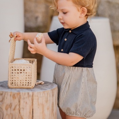 Criança de macacão azul escuro com parte inferior riscada a segurar conchas de caixa de rattan em tronco de madeira.