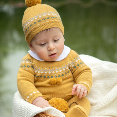 Bebé com roupa de malha amarela e gorro com pompom sentado numa cadeira com manta branca