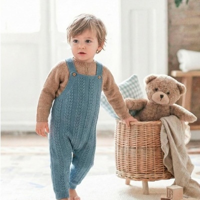 Menino pequeno com jardineira azul de lã e camiseta castanha num quarto claro com tapete e cesto de vime