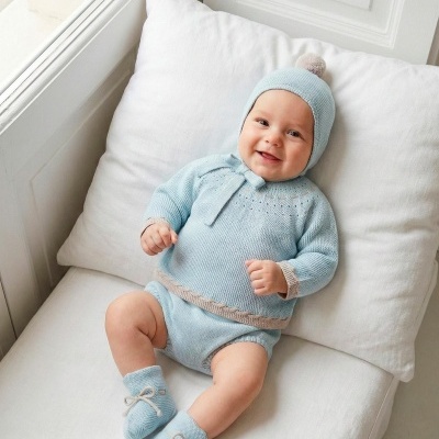 Bebé sorridente com roupa de malha azul claro e bege sentado num sofá branco.