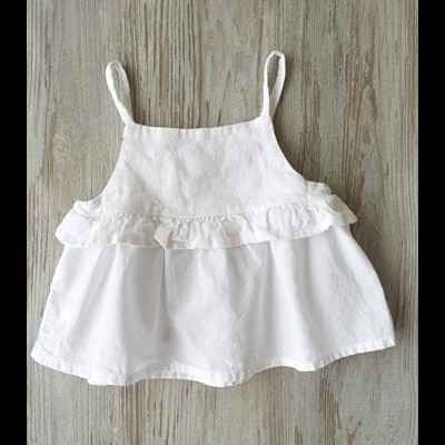 Top branco para criança com alças e folho, visto de frente sobre superfície de madeira
