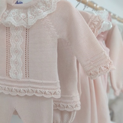 Conjunto de roupa de bebé rosa claro com gola de renda branca bordada e detalhes em croché pendurado em cabide de madeira.