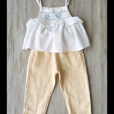 Conjunto infantil com blusa branca bordada azul e calças bege sobre piso de madeira clara