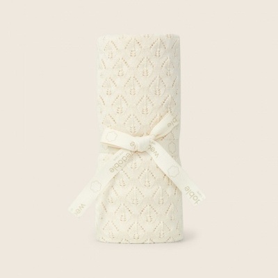 Manta de bebé creme em malha com padrão de folhas enrolada e presa com fita bege.