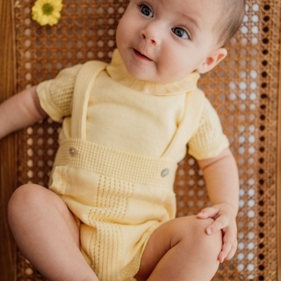 Bebé com roupa de malha amarela sentado numa cadeira de madeira e rattan com flor amarela ao lado.