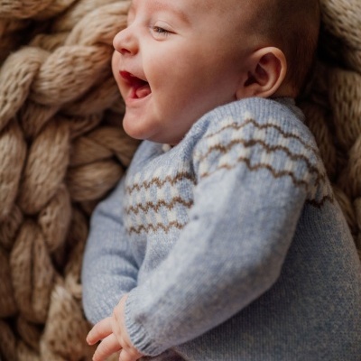 Bebé sorridente deitado em manta grossa bege com camisola cinza claro de malha com padrão castanho nas costas.