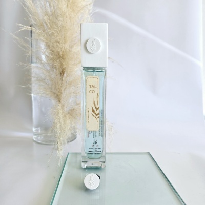 Frasco de perfume TAL CO em vidro com tampas brancas e etiqueta bege, sobre base de vidro e planta seca atrás.