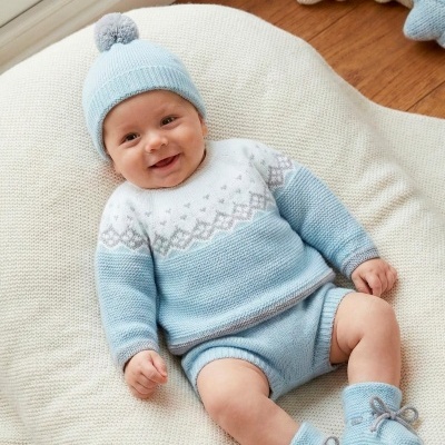 Bebé com roupa e gorro de malha azul e branca, deitado numa almofada creme
