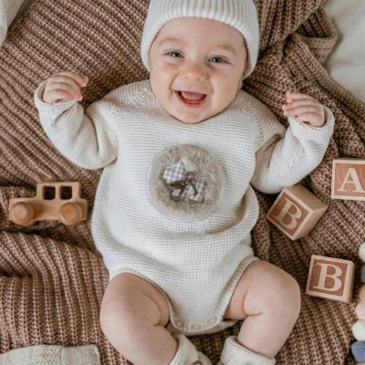 Bebé sorridente com roupa de malha branca e acessórios sobre manta castanha com blocos de madeira