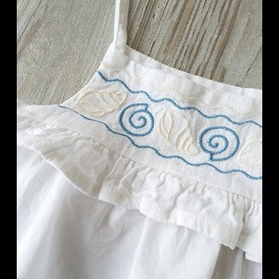 Parte superior de roupa branca com bordado azul e bege, alças finas, fundo madeira cinza