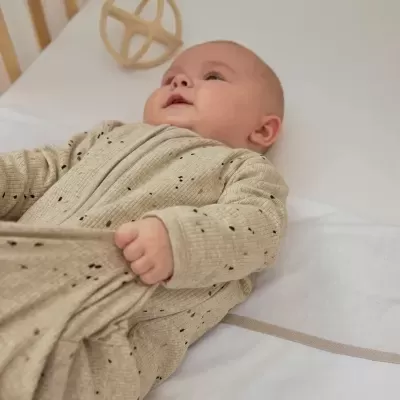Bebé deitado com roupa bege com pontos pretos num berço branco com brinquedo de madeira.