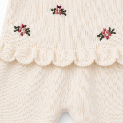 Roupa de criança em malha creme com bordados florais coloridos e folho decorativo.