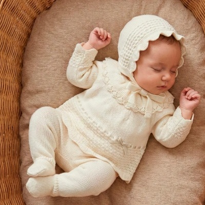 Bebé a dormir numa almofada bege dentro de um cesto de vime com roupa de malha creme.