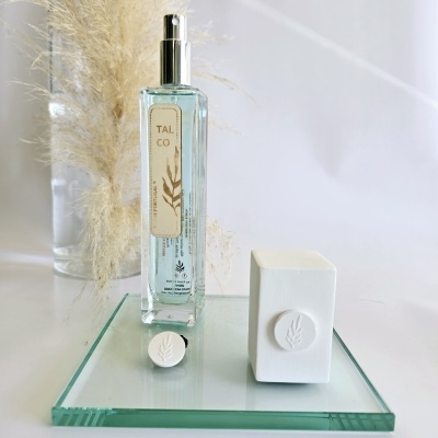 Frasco spray de vidro com líquido azul claro e rótulo Talco, tampa e objeto branco decorativo em base espelhada