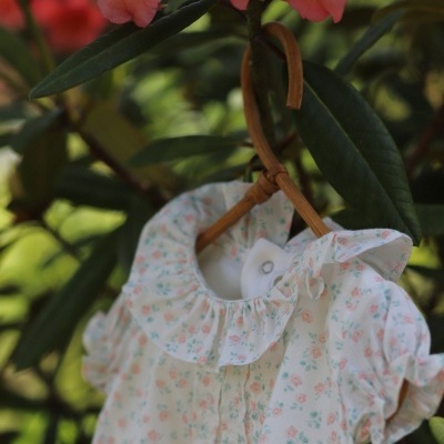 Vestido de bebé floral branco com gola frisada pendurado em cabide de madeira entre folhagem verde e flores cor-de-rosa