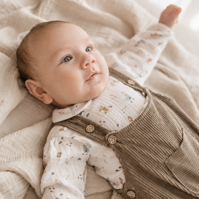 Bebé vestido com camisa branca estampada e jardineira castanha de veludo cotelê deitado sobre manta bege.