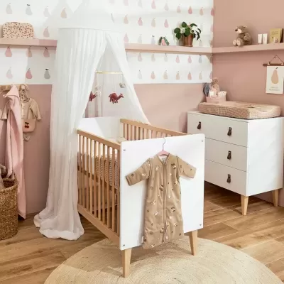 Quarto de bebé com berço de madeira clara e branca e mobília branca com puxadores em pele.