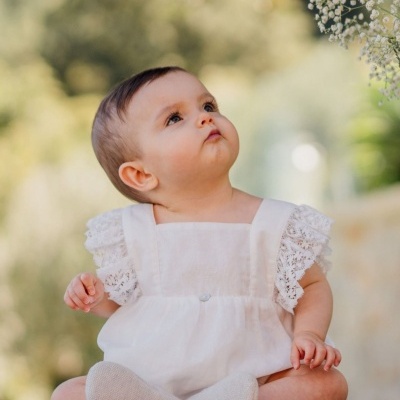 Bebé sentado em vestido branco com mangas rendadas e sapatinhos cinzentos ao ar livre