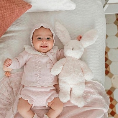 Bebé com roupa de malha rosa e ursa de pelúcia branca em cama branca