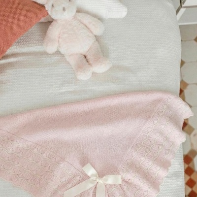 Manta de bebé rosa com rendas e laço sobre cama branca com peluche coelho e almofadas.