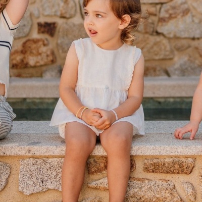 Criança com vestido branco e sapatos brancos sentada numa parede de pedra