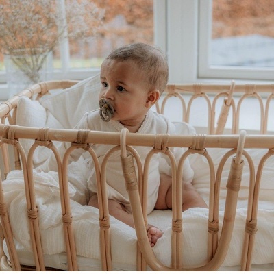 Berço de vime natural claro com bebé sentado em colchão branco perto de janela