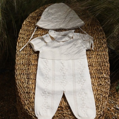Macacão e chapéu de bebé creme em tecido malhado com padrão e botões, sobre cadeira de fibras naturais