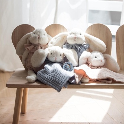 Quatro peluches coelhos em tecido macio sentados num banco de madeira clara perto de cortinas brancas