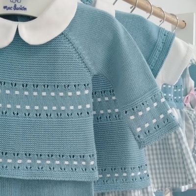 Conjunto de roupa de bebé azul de malha com gola branca pendurado em cabide de madeira.