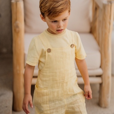 Criança com macacão curto amarelo claro e t-shirt amarela, cadeira de madeira ao fundo