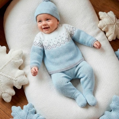 Bebé deitado com roupa de malha azul e branca numa almofada creme junto a almofadas de folhas azuis e creme