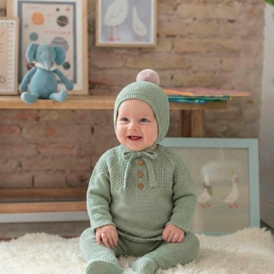 Bebé sentado com fato de malha verde claro e gorro com pompom, sentado em tapete branco felpudo com parede de tijolo e decoração infantil ao fundo