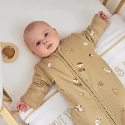 Bebé com saco de dormir castanho claro com desenhos de animais e folhas numa cama branca.