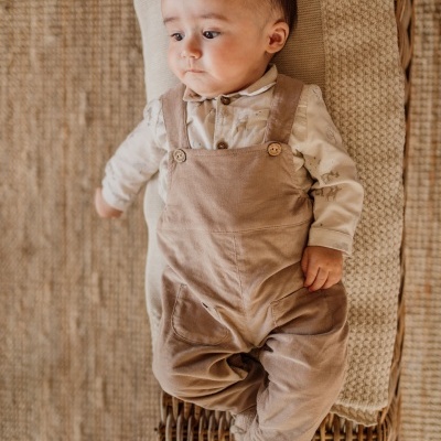 Bebé vestido com camisa creme estampada e calças camel de alças deitado em móvel de verga com colchão bege.