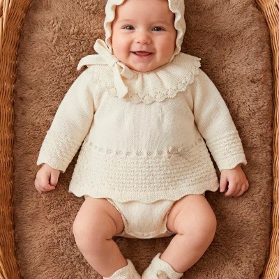 Bebé com roupa de malha creme deitada num cesto de vime com almofada castanha.