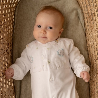 Bebé com macacão branco numa cama de verga castanha com almofada cinzenta