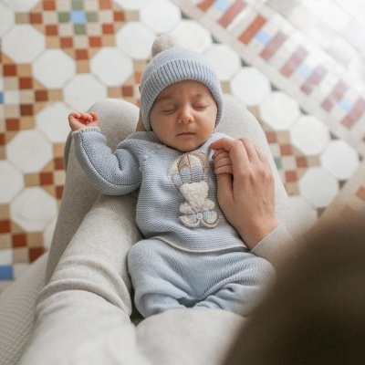 Bebé com conjunto de malha azul e boné, imagem de ursinho, em chão de azulejos coloridos, mãos de adulto a segurá-lo.
