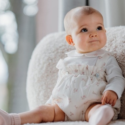 Bebé sentado em sofá branco com roupa branca e flores cor-de-rosa