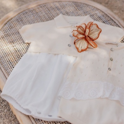 Roupa de bebé em creme com vestido e cardigan decorado com borboleta em palha sobre cesto de vime.